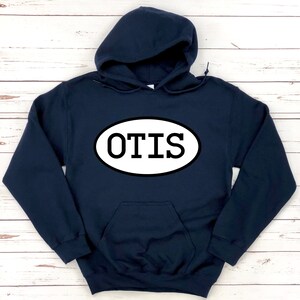 Op de afbeelding: Een marineblauwe hoodie met een voorzak en een witte ovale afbeelding met het woord "OTIS" in vette, zwarte letters. De hoodie heeft een capuchon met trekkoord en lange mouwen.