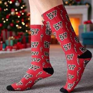 May include: Red socks with a repeating pattern of the number "57" in green and white. The socks have black heels and toes. The background includes a Christmas tree and a fireplace.