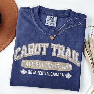 May include: A blue short-sleeved t-shirt with a distressed beige graphic that reads CABOT TRAIL, CAPE BRETON ISLAND, and NOVA SCOTIA, CANADA, featuring two maple leaf icons. The shirt has a Comfort Colors brand label showing size S.