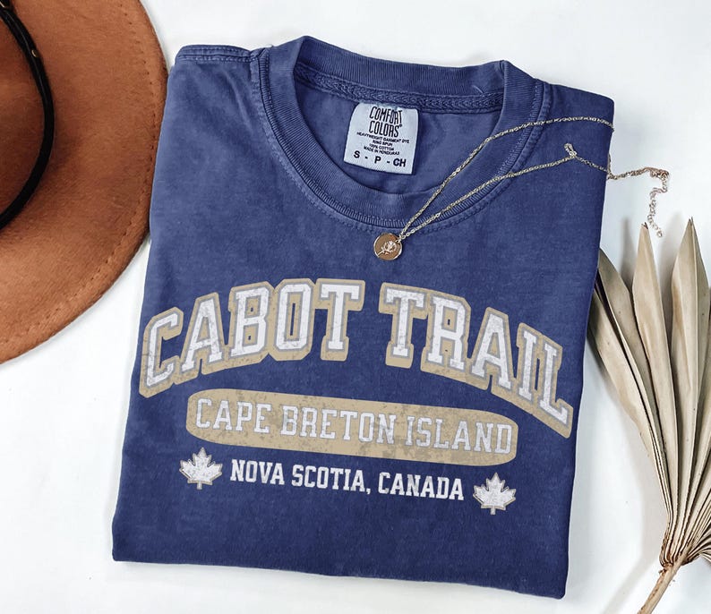 May include: A blue short-sleeved t-shirt with a distressed beige graphic that reads CABOT TRAIL, CAPE BRETON ISLAND, and NOVA SCOTIA, CANADA, featuring two maple leaf icons. The shirt has a Comfort Colors brand label showing size S.