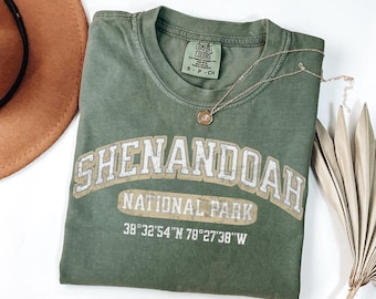 May include: An olive green crew neck t-shirt featuring the white and tan text SHENANDOAH NATIONAL PARK and the coordinates 38°32'54"N 78°27'38"W. The shirt has a Comfort Colors brand label on the inside collar indicating a size small.