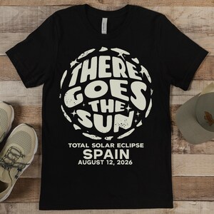 May include: Black t-shirt with a white graphic that reads "THERE GOES THE SUN" in a circular design. Below the graphic, the text reads "TOTAL SOLAR ECLIPSE SPAIN AUGUST 12, 2026".