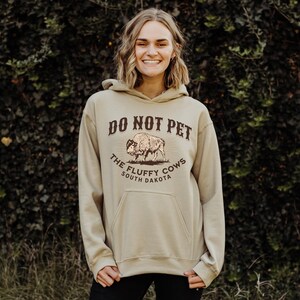 May include: A tan hoodie with a brown graphic that says "Do Not Pet The Fluffy Cows South Dakota" and a drawing of a bison.