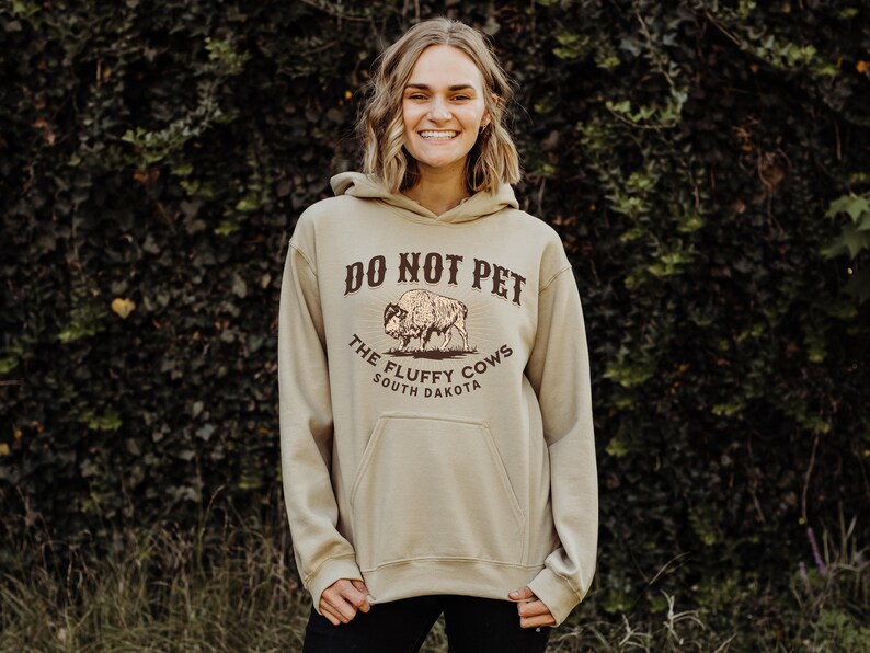 May include: A tan hoodie with a brown graphic that says "Do Not Pet The Fluffy Cows South Dakota" and a drawing of a bison.
