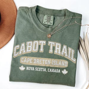 May include: An olive green short-sleeve t-shirt with a crew neckline. The front features white distressed text reading CABOT TRAIL, CAPE BRETON ISLAND, and NOVA SCOTIA, CANADA, flanked by two maple leaf icons. A Comfort Colors brand tag is visible.
