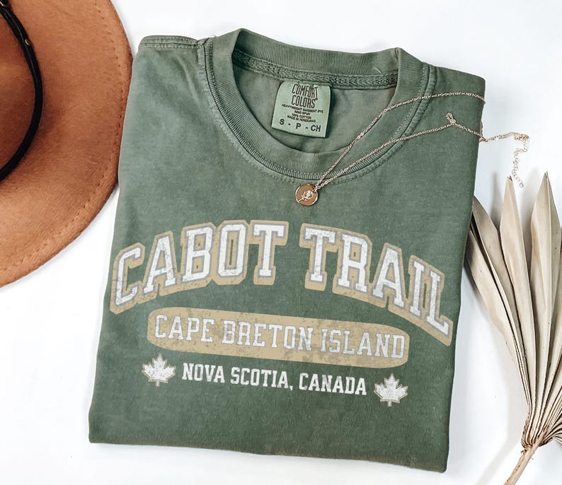 May include: An olive green short-sleeve t-shirt with a crew neckline. The front features white distressed text reading CABOT TRAIL, CAPE BRETON ISLAND, and NOVA SCOTIA, CANADA, flanked by two maple leaf icons. A Comfort Colors brand tag is visible.
