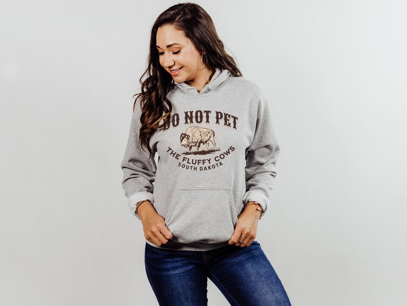 May include: A grey hooded sweatshirt with a brown graphic print of a buffalo and the text "Do Not Pet The Fluffy Cows South Dakota".