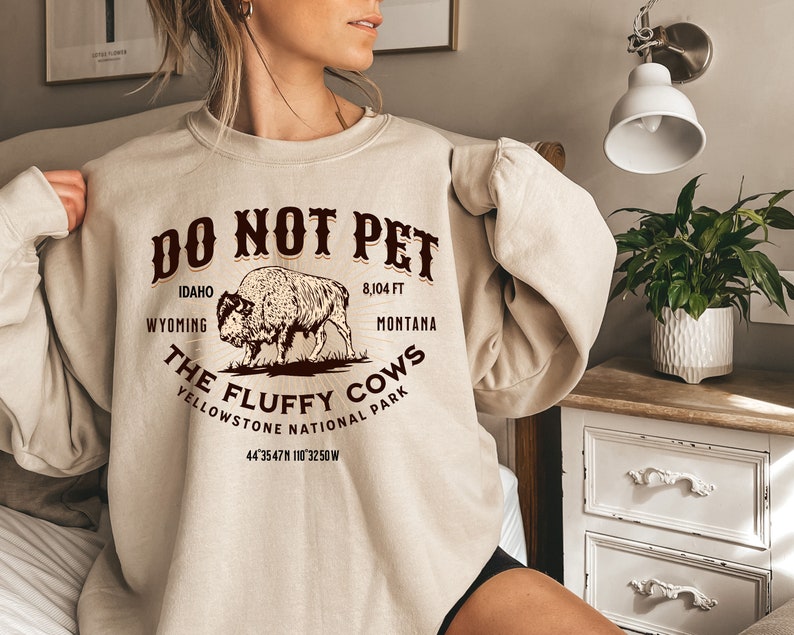 May include: A beige sweatshirt with a brown graphic print that says "Do Not Pet The Fluffy Cows Yellowstone National Park 44'3547N 110'3250W". The graphic also includes a drawing of a bison.