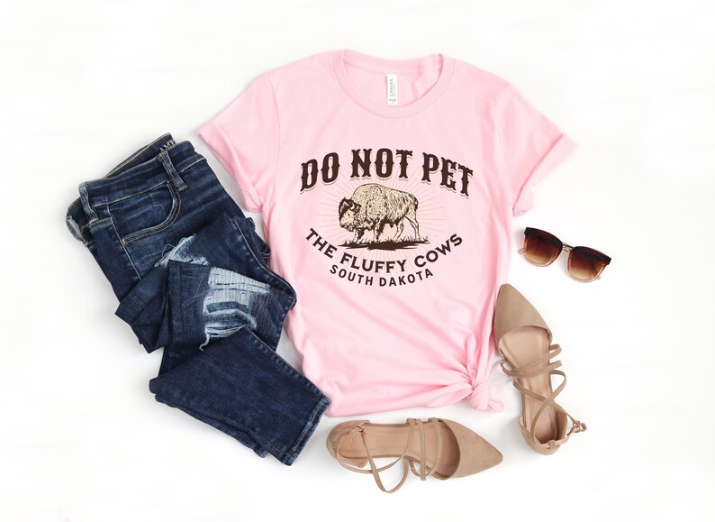 Peut inclure: T-shirt rose avec un graphique brun d'un bison et le texte "Do Not Pet The Fluffy Cows South Dakota".