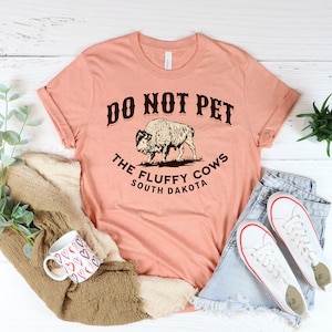 Peut inclure: Un t-shirt couleur p&ecirc;che avec le texte "Do Not Pet The Fluffy Cows South Dakota" et un dessin de bison.