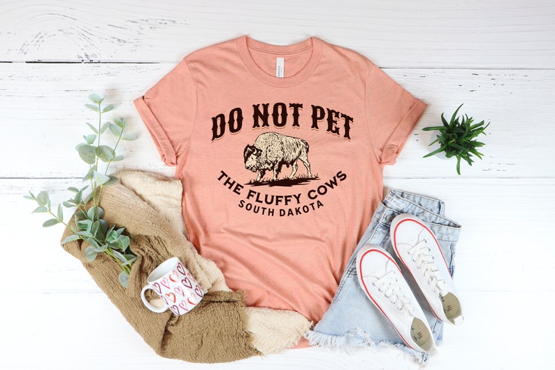 Peut inclure: Un t-shirt couleur p&ecirc;che avec le texte "Do Not Pet The Fluffy Cows South Dakota" et un dessin de bison.