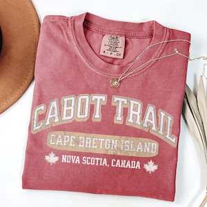 May include: A dusty rose short-sleeve t-shirt featuring the text CABOT TRAIL, CAPE BRETON ISLAND, and NOVA SCOTIA, CANADA in a distressed white and tan font. The graphic includes two maple leaf icons. The shirt has a Comfort Colors brand label.