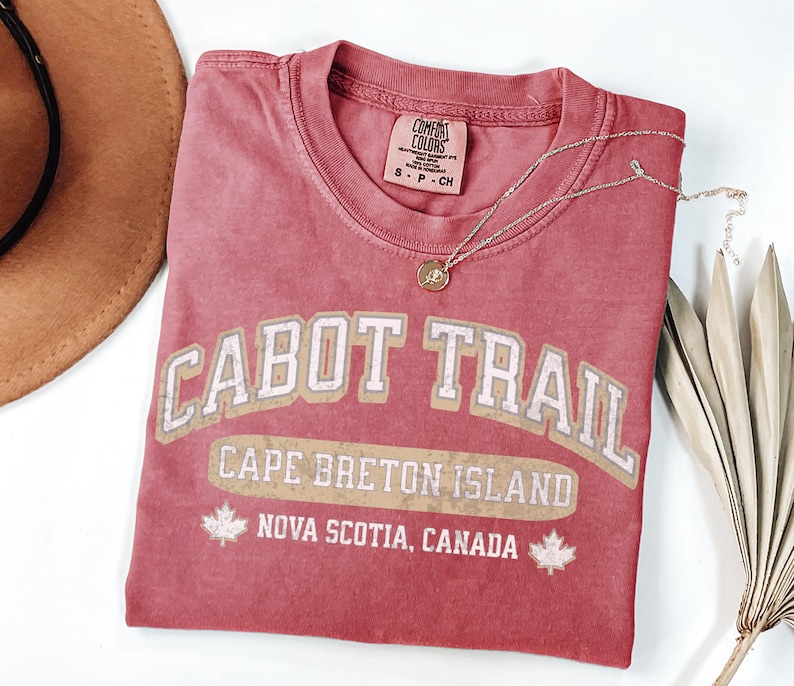 May include: A dusty rose short-sleeve t-shirt featuring the text CABOT TRAIL, CAPE BRETON ISLAND, and NOVA SCOTIA, CANADA in a distressed white and tan font. The graphic includes two maple leaf icons. The shirt has a Comfort Colors brand label.