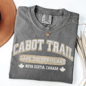 May include: A charcoal grey short-sleeve t-shirt featuring the text CABOT TRAIL, CAPE BRETON ISLAND, and NOVA SCOTIA, CANADA in a distressed white and tan font. Two small maple leaf icons flank the bottom text. The shirt has a Comfort Colors label.