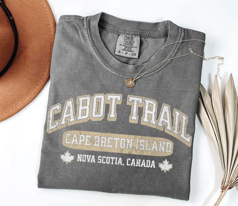 May include: A charcoal grey short-sleeve t-shirt featuring the text CABOT TRAIL, CAPE BRETON ISLAND, and NOVA SCOTIA, CANADA in a distressed white and tan font. Two small maple leaf icons flank the bottom text. The shirt has a Comfort Colors label.