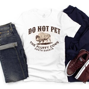 Peut inclure: T-shirt blanc avec un graphique brun qui dit "Do Not Pet The Fluffy Cows South Dakota". Le graphique repr&eacute;sente un bison brun.