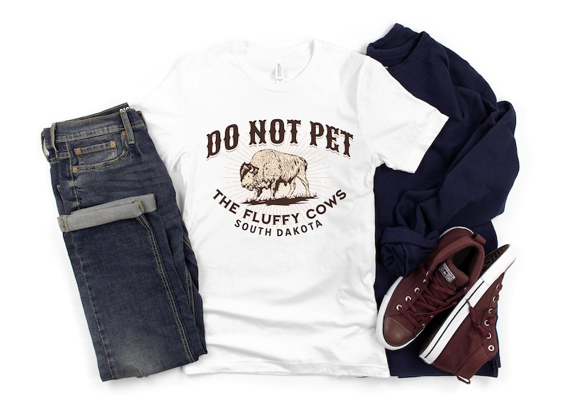 Peut inclure: T-shirt blanc avec un graphique brun qui dit "Do Not Pet The Fluffy Cows South Dakota". Le graphique repr&eacute;sente un bison brun.