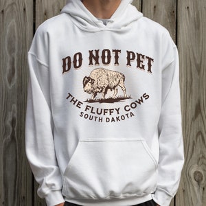 May include: White hooded sweatshirt with a brown graphic that says "Do Not Pet The Fluffy Cows South Dakota" and a drawing of a bison.