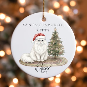May include: A white ceramic Christmas ornament featuring a fluffy white cat wearing a Santa hat, sitting next to a decorated Christmas tree. The ornament has the text "SANTA'S FAVORITE KITTY" and the name "Oscar" with the year "2025".