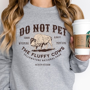 May include: Gray sweatshirt with a brown graphic print that says "Do Not Pet The Fluffy Cows Yellowstone National Park" with a bison illustration and the text "Idaho, Wyoming, 8,104 ft, Montana, 44&deg;35'47"N 110&deg;32'50"W".