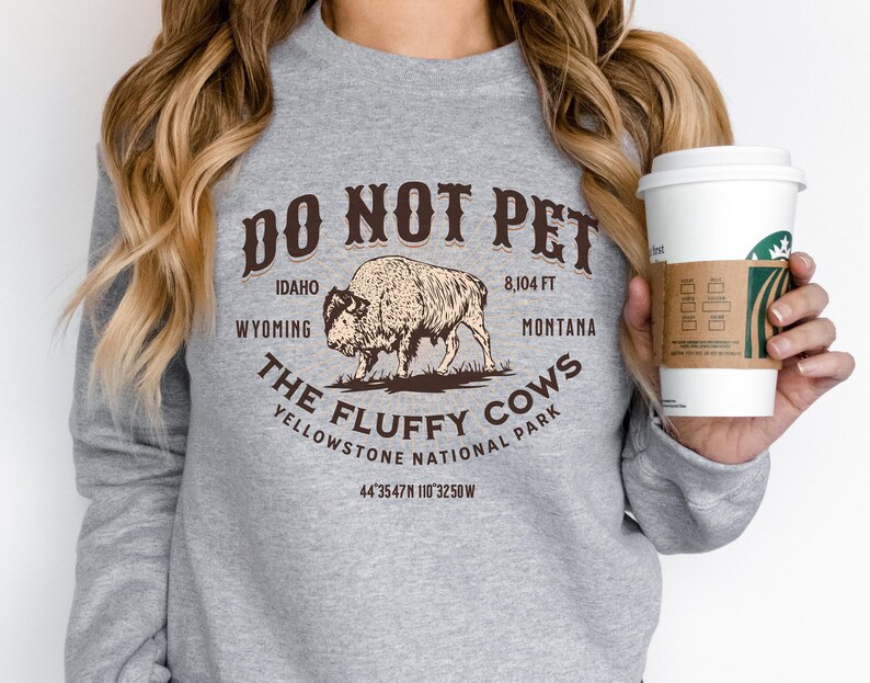 May include: Gray sweatshirt with a brown graphic print that says "Do Not Pet The Fluffy Cows Yellowstone National Park" with a bison illustration and the text "Idaho, Wyoming, 8,104 ft, Montana, 44&deg;35'47"N 110&deg;32'50"W".