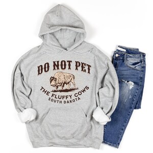 May include: A grey hooded sweatshirt with the text "Do Not Pet The Fluffy Cows South Dakota" and a brown illustration of a bison.