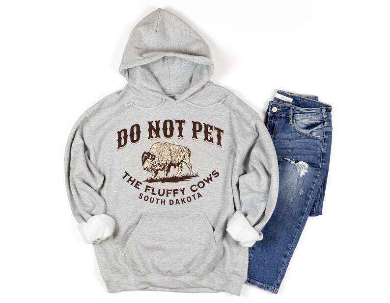 May include: A grey hooded sweatshirt with the text "Do Not Pet The Fluffy Cows South Dakota" and a brown illustration of a bison.