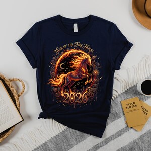May include: Navy blue t-shirt with a fiery horse design and the text "Year of the Fire Horse 2026". The graphic includes a horse within a circular frame with celestial elements. The shirt is folded on a patterned blanket.