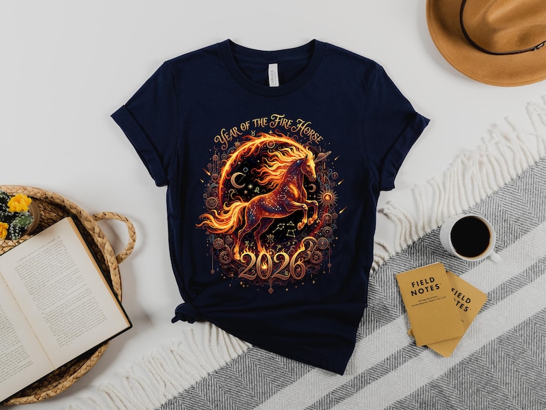 May include: Navy blue t-shirt with a fiery horse design and the text "Year of the Fire Horse 2026". The graphic includes a horse within a circular frame with celestial elements. The shirt is folded on a patterned blanket.