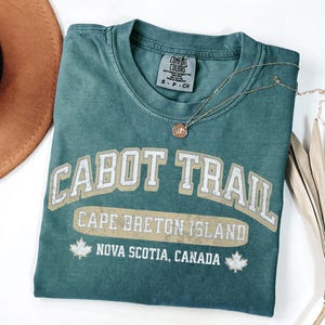 May include: A teal short-sleeved t-shirt with a crew neckline featuring the text CABOT TRAIL, CAPE BRETON ISLAND, and NOVA SCOTIA, CANADA in white and beige lettering, accompanied by two small white maple leaf icons on the lower graphic.