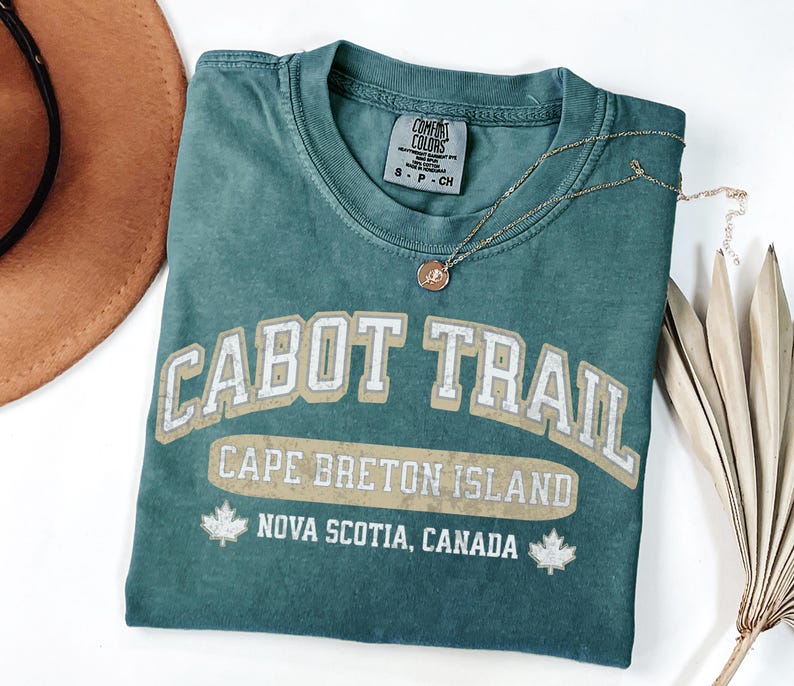May include: A teal short-sleeved t-shirt with a crew neckline featuring the text CABOT TRAIL, CAPE BRETON ISLAND, and NOVA SCOTIA, CANADA in white and beige lettering, accompanied by two small white maple leaf icons on the lower graphic.