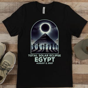 Pode incluir: Camiseta preta com um gráfico de um eclipse solar sobre uma pirâmide com silhuetas de pessoas na frente dela. O texto "Total Solar Eclipse Egypt August 2, 2027" é impresso abaixo do gráfico.