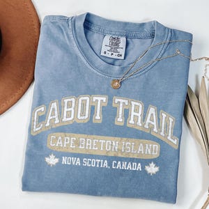 May include: A blue short-sleeve t-shirt with a distressed graphic that reads CABOT TRAIL, CAPE BRETON ISLAND, and NOVA SCOTIA, CANADA, featuring two maple leaf icons. The shirt has a Comfort Colors brand label on the inside collar and a size S tag.