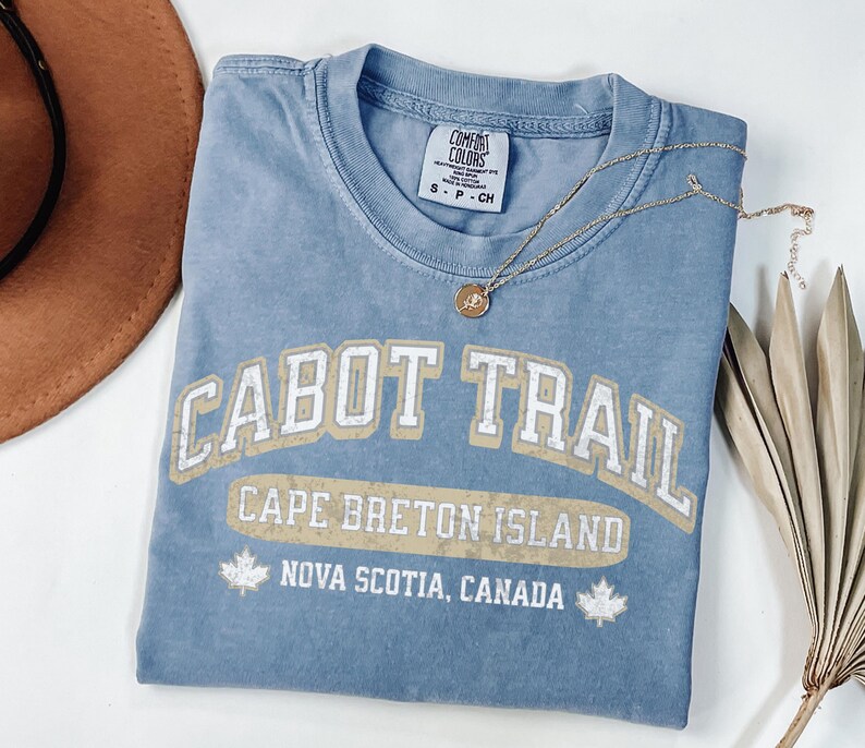 May include: A blue short-sleeve t-shirt with a distressed graphic that reads CABOT TRAIL, CAPE BRETON ISLAND, and NOVA SCOTIA, CANADA, featuring two maple leaf icons. The shirt has a Comfort Colors brand label on the inside collar and a size S tag.