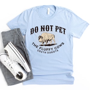 Peut inclure: T-shirt bleu clair avec un graphique brun d'un bison et le texte "Do Not Pet The Fluffy Cows South Dakota".
