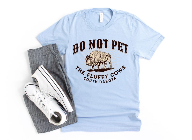 Peut inclure: T-shirt bleu clair avec un graphique brun d'un bison et le texte "Do Not Pet The Fluffy Cows South Dakota".