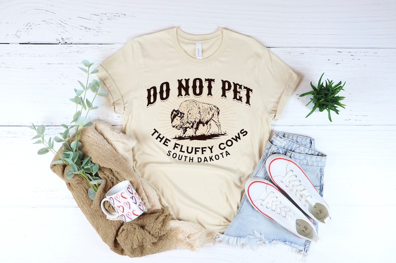 Peut inclure: Un t-shirt beige avec un graphique brun d'un bison et le texte "Do Not Pet The Fluffy Cows South Dakota".