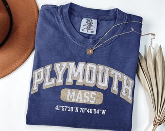 May include: A blue short-sleeve t-shirt with a crew neckline. The front features the text PLYMOUTH MASS in a distressed white and beige font, with the coordinates 41°57'30"N 70°40'04"W printed below. A small Comfort Colors brand tag is at the neck.
