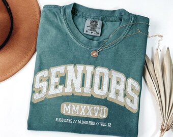 May include: A teal short-sleeve t-shirt featuring the white text SENIORS in a collegiate font, the Roman numeral MMXXVII, and the text 2,160 DAYS // 14,040 HRS // VOL. 12. The shirt has a Comfort Colors brand label at the inner neckline.