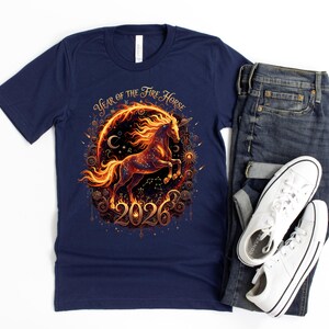 May include: A navy blue t-shirt with a fiery horse graphic, encircled by a decorative frame, and the text "Year of the Fire Horse 2026". The shirt is styled with blue jeans and white trainers for a casual look.