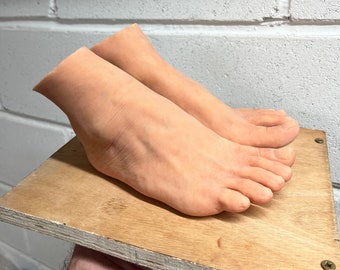 May include: A pair of realistic-looking silicone feet, with toes and a natural skin tone, are displayed on a wooden surface.