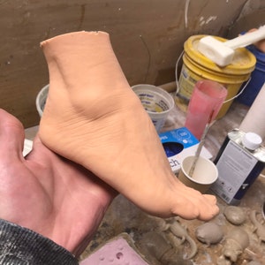 Op de afbeelding: Een close-up van een vleeskleurige voet die van klei is gemaakt. De voet wordt vastgehouden door een hand en rust op een houten oppervlak. Er zijn andere kleisculpturen op de achtergrond.