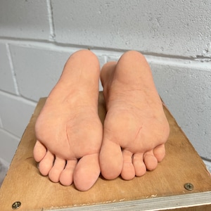 May include: Two bare feet are placed on a wooden surface. The feet are positioned side by side with the toes pointing towards the camera. The feet are light pink in color and have visible lines and wrinkles.