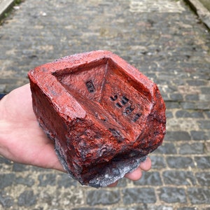 May include: A red brick with a rough, textured surface. The brick has a small, rectangular indentation on the top and a larger, rectangular indentation on the side. The brick is covered in a layer of red paint.
