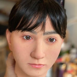 Pode incluir: Uma cabeça de silicone realista com cabelo curto e escuro e olhos castanhos. O rosto tem um tom de pele natural com sardas sutis e lábios rosados. A cabeça é segurada por uma mão, com um fundo desfocado.