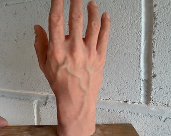 May include: A realistic-looking, life-size, silicone hand model with visible veins. The hand is posed with the fingers spread apart and the palm facing up. The hand is resting on a wooden board.