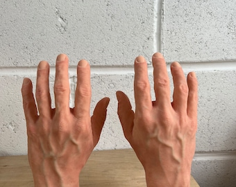 May include: Two realistic-looking hands with detailed veins, made of a flesh-toned material, are displayed on a wooden base. The hands are positioned with palms facing outward, fingers slightly spread.