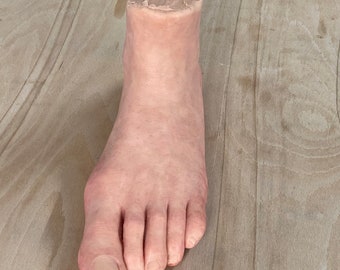 May include: A realistic-looking prosthetic foot made of silicone, with a flesh-toned color and a smooth surface. The foot is missing the toes and part of the ankle, and the cut edge is visible.
