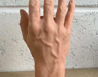 May include: A realistic-looking, detailed, flesh-toned silicone hand model with visible veins. The hand is posed with fingers spread apart and is resting on a wooden surface.