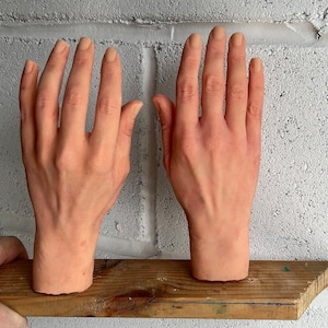 Pode incluir: Duas mãos de silicone de aparência realista, cor de carne, com escultura detalhada dos dedos e unhas. As mãos estão posicionadas em uma superfície de madeira.